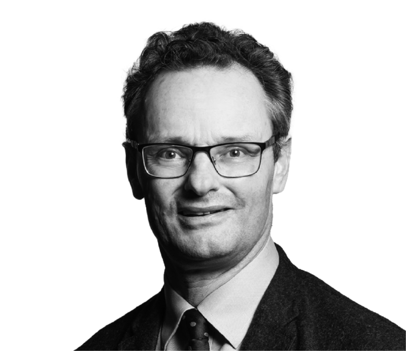 A man with curly hair and glasses smiles for the camera, wearing a dark suit jacket and light-colored shirt. The background is plain white.