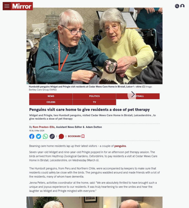 Two elderly women at a care home hold a small penguin. One woman smiles while the other looks at the penguin with interest. The scene captures a heartwarming moment during a visit from penguins to the residents.