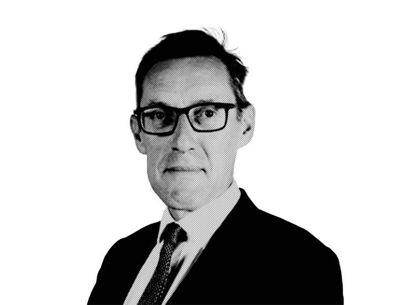 A man in a suit and tie, wearing glasses, poses against a white background. His expression is serious, and his hair is slightly tousled.