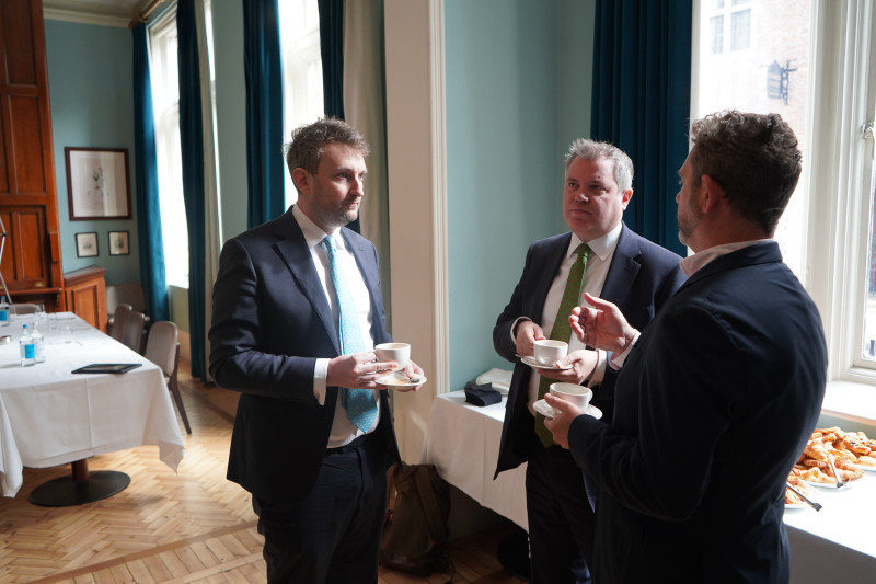 Three men in business attire engage in conversation while holding cups of coffee. They stand in a well-lit room with large windows and blue drapes, surrounded by plates of food on a nearby table.
