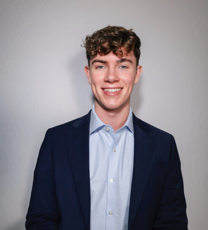 A young man with curly hair smiles confidently while wearing a dark blazer over a light blue shirt. The background is plain and light-colored.