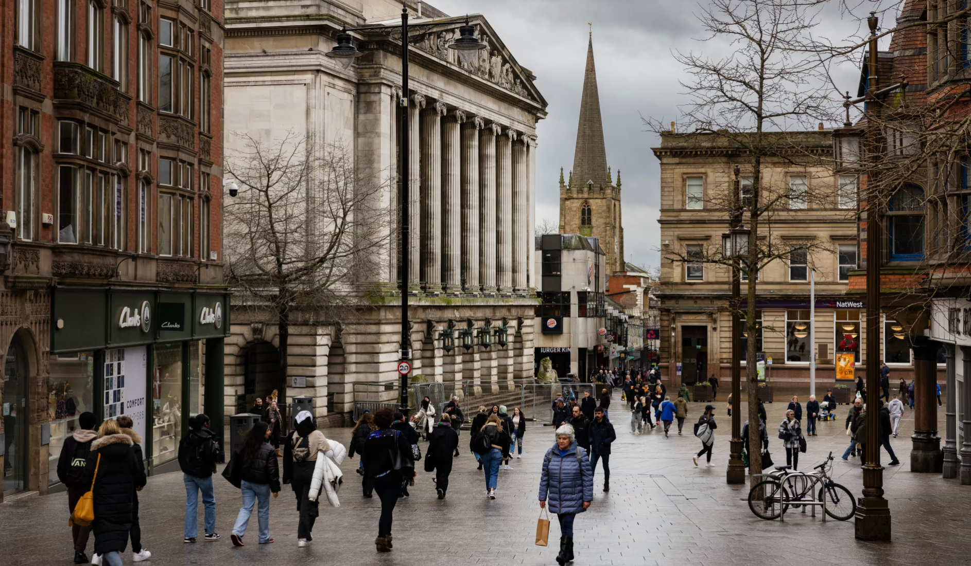 A bustling city street with pedestrians walking along the pavement, lined with shops and historic buildings. In the background, a church spire reaches towards a cloudy sky, contrasting with the modern architecture nearby.