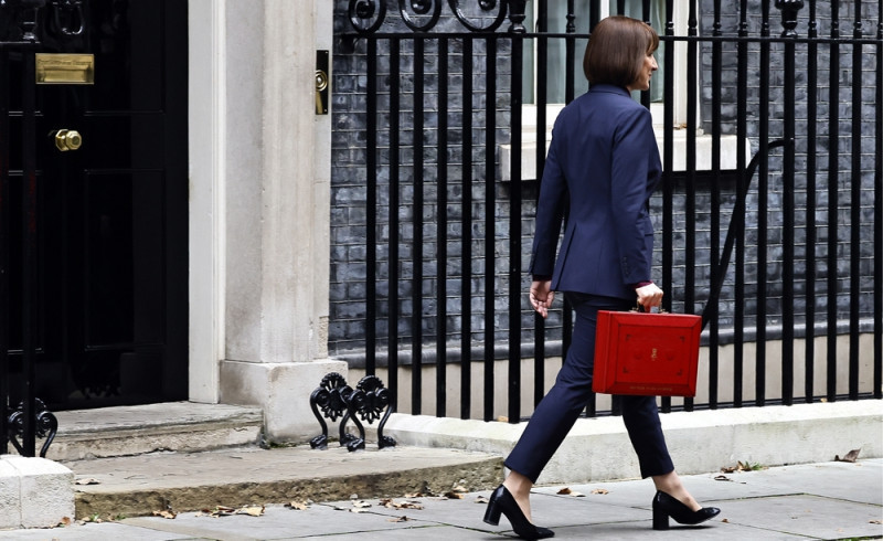 A woman in a tailored navy suit walks confidently past a black wrought-iron gate, carrying a bright red briefcase.