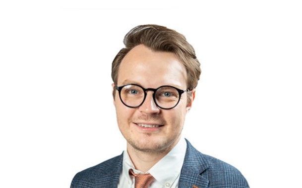 A man with glasses and a light blue suit jacket smiles confidently. He has neat hair styled to the side and is wearing a white shirt with a tie.