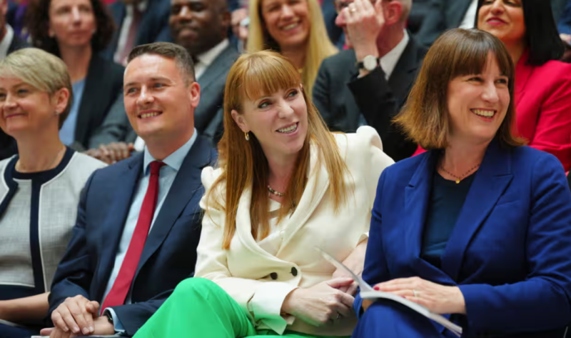 A group of smiling people seated together, engaged and attentive during an event. One woman has long red hair and is wearing a white blazer with green pants, while another woman in a navy suit sits beside her, both appearing joyful. A man in a suit smiles warmly in the foreground.