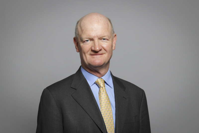 A man with a bald head and a slight smile is wearing a dark suit and a light-colored tie, posing against a plain gray background.