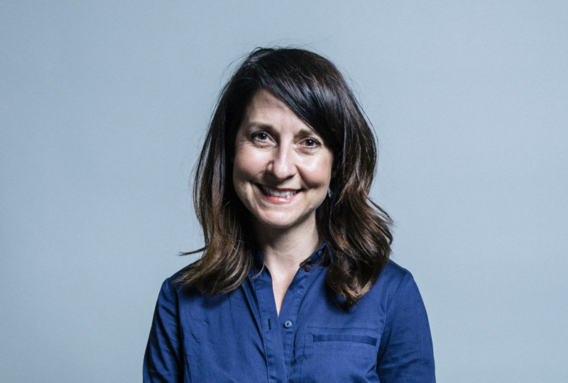 A woman with long, dark hair smiles at the camera while wearing a blue shirt against a light gray background.