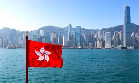 The red flag of Hong Kong flies in the foreground, with a panoramic view of the Hong Kong skyline featuring skyscrapers and mountains in the background.