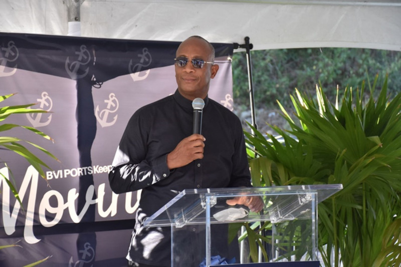 A man in a black shirt stands behind a clear podium, speaking at an event. He smiles confidently while addressing an audience, with a backdrop featuring a logo and the word "Moving." Green plants are visible in the foreground.