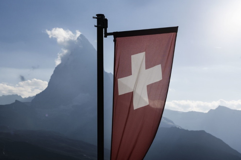 A Swiss flag with a white cross stands prominently in the foreground, with the Matterhorn mountain in the background under a cloudy sky.