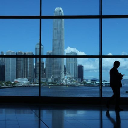 A silhouette of a person standing indoors, viewing a vibrant city skyline with a tall skyscraper and a Ferris wheel near the water. The scene is framed by large windows, showcasing a clear blue sky and fluffy white clouds.