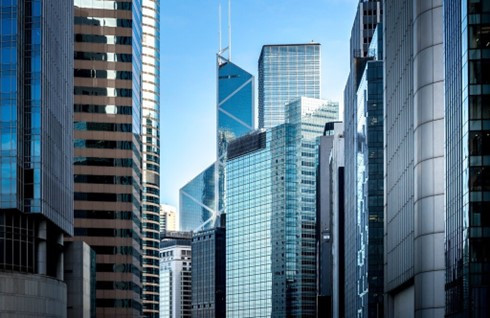 A view of skyscrapers with reflective glass surfaces against a clear blue sky. The buildings feature a mix of modern architectural designs, including sharp angles and sleek lines.