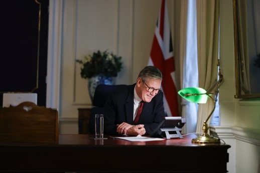 A man sitting at a wooden desk, smiling while looking at a phone. He is in a room with a British flag in the background and a green desk lamp illuminating the workspace. A glass of water is placed beside him.