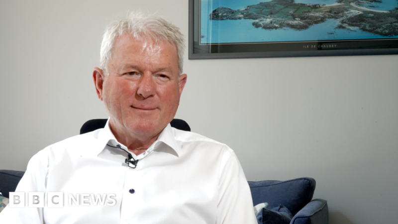 A man with short gray hair wearing a white button-up shirt sits in a well-lit room, smiling at the camera. A coastal landscape artwork is visible in the background.