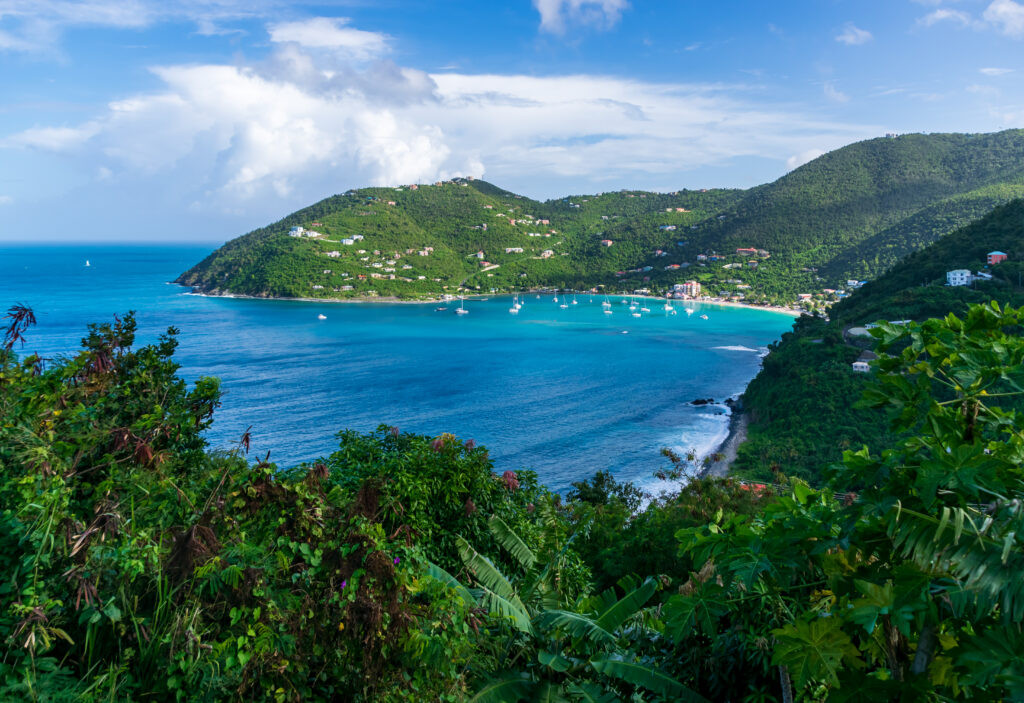 A scenic view of a bay surrounded by lush green hills and calm turquoise waters, with boats anchored near the shore and a partly cloudy sky.