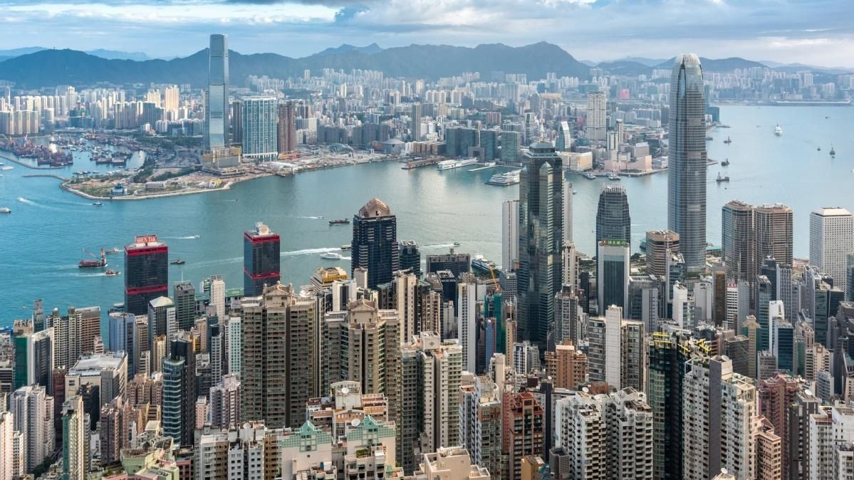 A panoramic view of Hong Kong's skyline, featuring a dense cluster of skyscrapers, with Victoria Harbour in the foreground and mountainous terrain in the background.