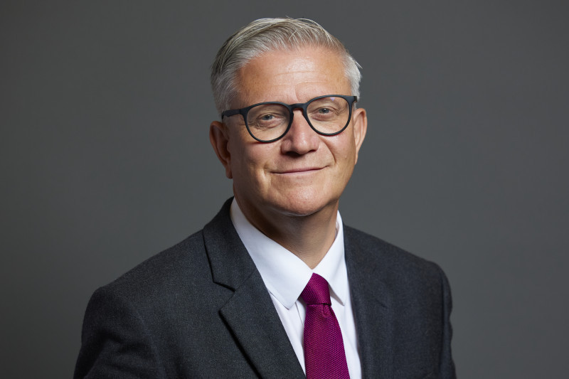 A middle-aged man with gray hair and glasses wears a dark suit and a bright pink tie, smiling confidently against a neutral gray background.