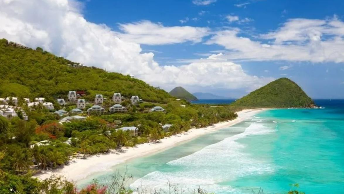 A scenic view of a tropical beach with white sand and turquoise waters, framed by lush green hills and scattered resort buildings. White fluffy clouds dot the blue sky, creating a serene atmosphere.