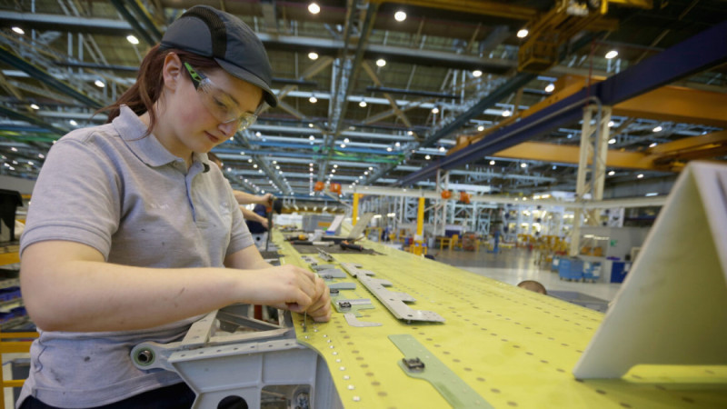 A woman in safety glasses and a cap works on assembling an aircraft component at a manufacturing facility. She focuses intently on her task, surrounded by machinery and other workers in the background.