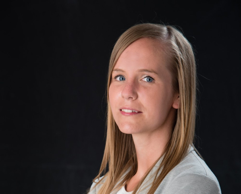 A young woman with long straight blonde hair and blue eyes is looking slightly to the side. She has a neutral expression and is wearing a light-colored top against a dark background.