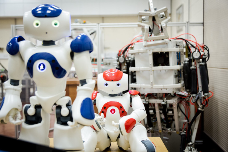 Two humanoid robots in a lab setting. The foreground features a blue and white robot with glowing eyes, while the background shows a larger, wire-framed robot with a skeletal appearance.