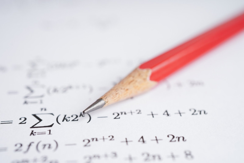 A close-up of a sharpened red pencil resting on a sheet of paper featuring mathematical equations and symbols.