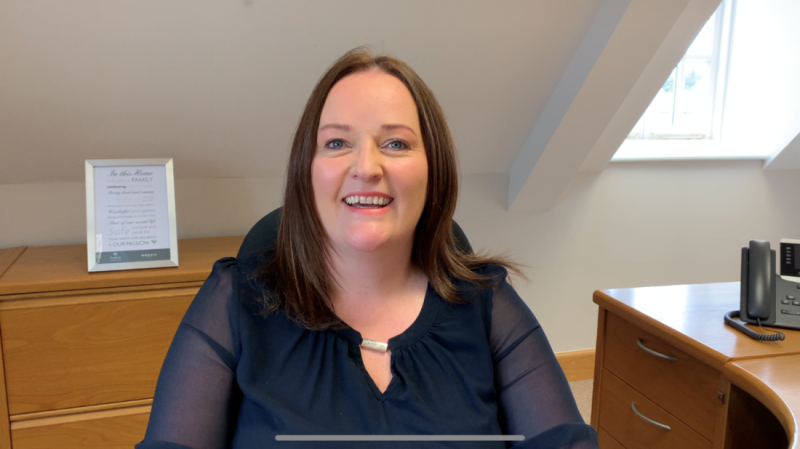 A woman with long brown hair is smiling while sitting at a desk in a well-lit office. She is wearing a navy blue blouse and is positioned in front of a wooden desk and a phone.