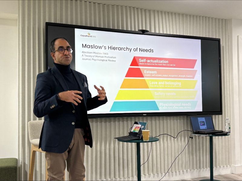 A speaker presenting on Maslow's Hierarchy of Needs, with a colorful chart displayed on a screen behind them. The speaker is wearing a blue blazer and glasses, gesturing towards the chart while engaging the audience.
