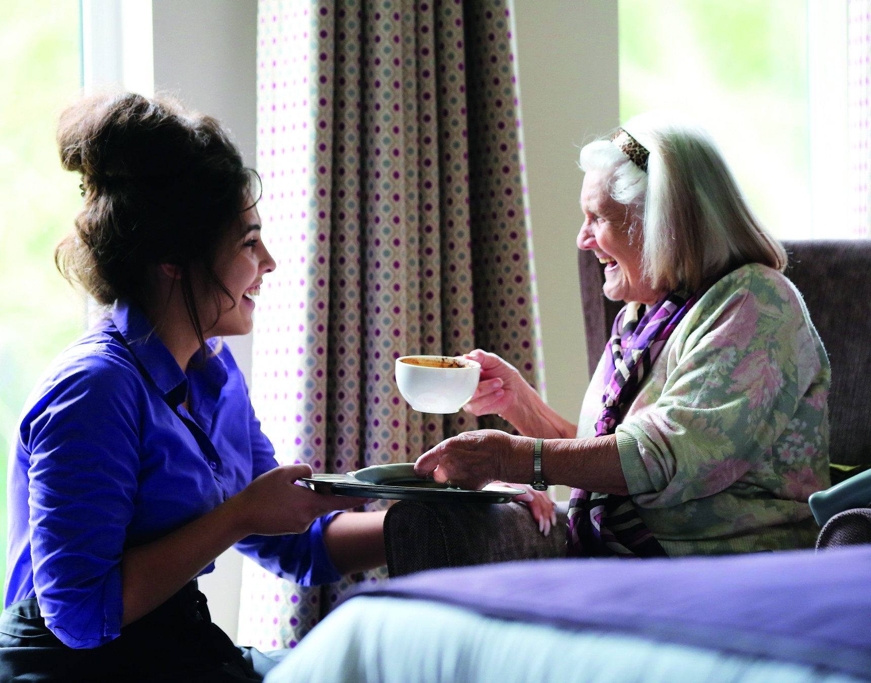 A caregiver smiling as she serves a cup of tea to an elderly woman sitting in a chair. The setting features light streaming through a window and patterned curtains.