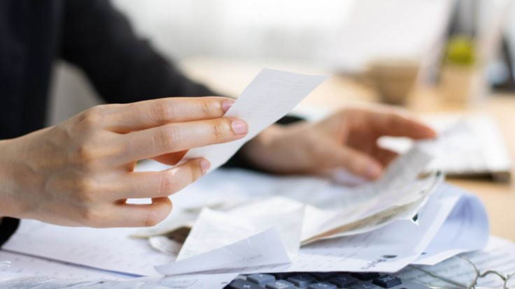 A person holds a receipt while examining a cluttered desk filled with papers and a calculator.