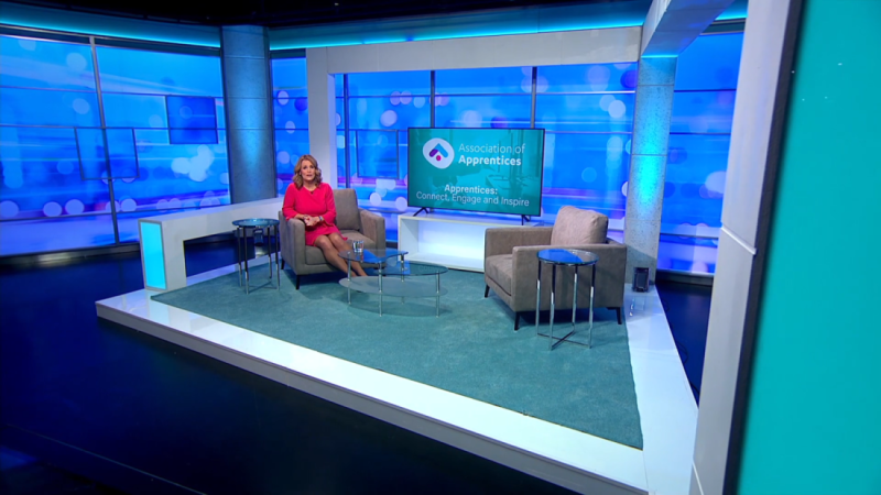 A well-lit television studio set featuring two beige armchairs and a round glass table on a teal rug. A backdrop with the logo of the Association of Apprentices is displayed, along with the text "Apprentices Connect, Engage and Inspire."