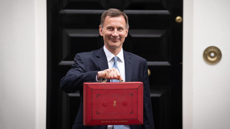 A man in a suit smiles while holding a red briefcase outside a black door with a brass doorknob.