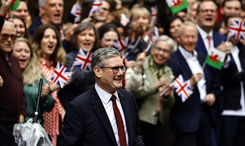 A man in a suit smiles as he walks through a crowd of enthusiastic supporters holding British and Welsh flags.