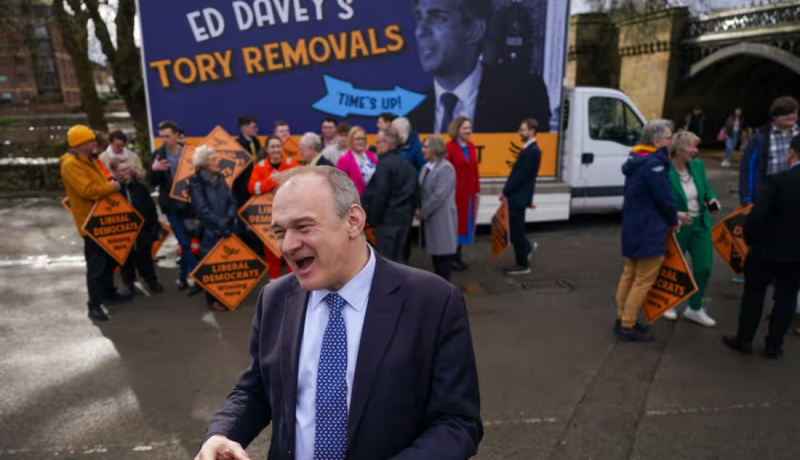 A group of people gathers near a campaign van displaying a large banner criticizing Ed Davey, with individuals holding signs for the Liberal Democrats. The scene is lively, with diverse attendees engaged in conversation and activities.