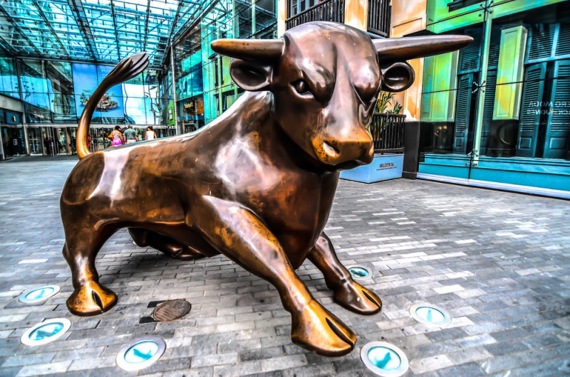 A large bronze bull sculpture positioned in an urban setting with glass architecture in the background. The bull is depicted in a crouching stance, with a shiny surface reflecting light.