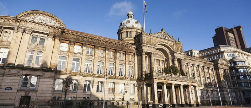 Victorian-style building with a grand façade, large columns, and a dome, under a blue sky. The structure features intricate architectural details and is flanked by modern buildings.