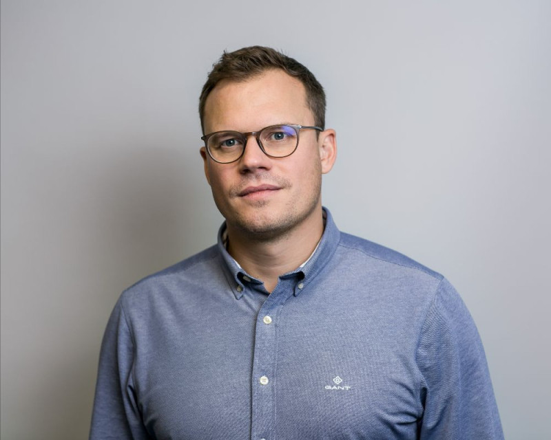 A man with short brown hair and glasses is standing against a light gray background. He is wearing a blue button-up shirt and looking directly at the camera with a neutral expression.