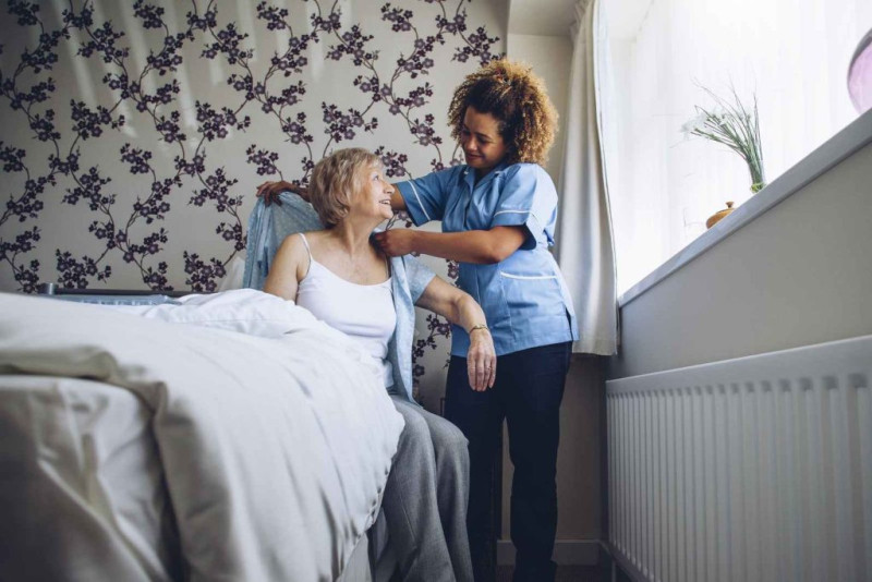 A caregiver assists an elderly woman in a bedroom setting, helping her put on a sweater. The room features floral wallpaper and a window with soft light.