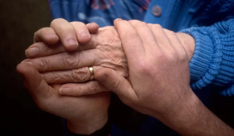 Two hands of different ages are clasped together, with one hand showing signs of aging and a wedding band. The other hand appears younger, gently holding the older hand. The contrasting skin textures highlight a connection across generations.