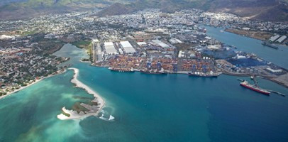 Aerial view of a coastal town featuring a busy port with shipping containers, waterfront facilities, and clear turquoise waters. Nearby, a sandy beach and lush green hills are visible in the background.