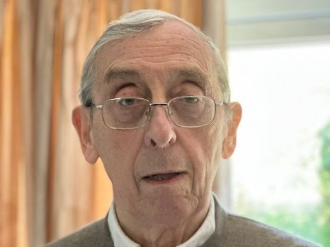An elderly man with gray hair and glasses stands indoors, looking directly at the camera with a neutral expression. He wears a light sweater over a collared shirt, with soft natural light illuminating the background.