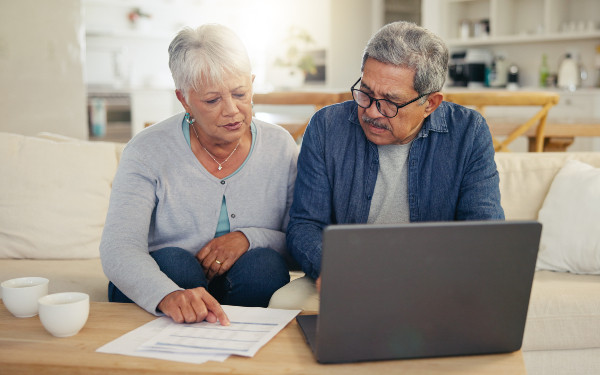 A senior couple sitting on a couch, looking at a laptop. The woman is pointing at a document in her hand while the man observes attentively. Two cups are placed on the coffee table.