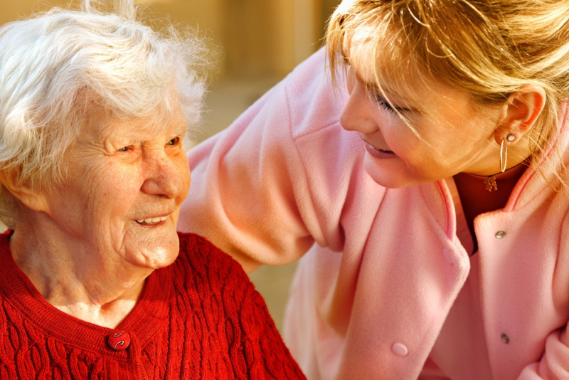 An elderly woman with white hair smiles while looking at a younger woman, who is wearing a pink jacket and leaning in, sharing a warm and caring moment.