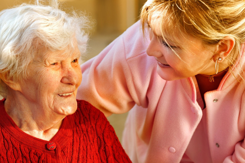 A smiling elderly woman with white hair sits closely beside a young woman, who is wearing a pink jacket and looking affectionately at her. Their warm interaction conveys a sense of companionship and care.