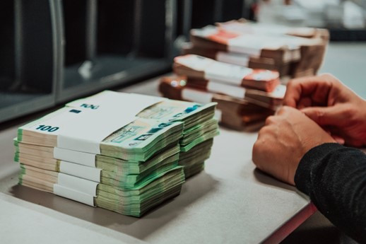 A stack of Canadian hundred-dollar bills is neatly arranged on a table, with additional stacks of different denominations in the background. A person's hands are seen sorting and handling the bills.