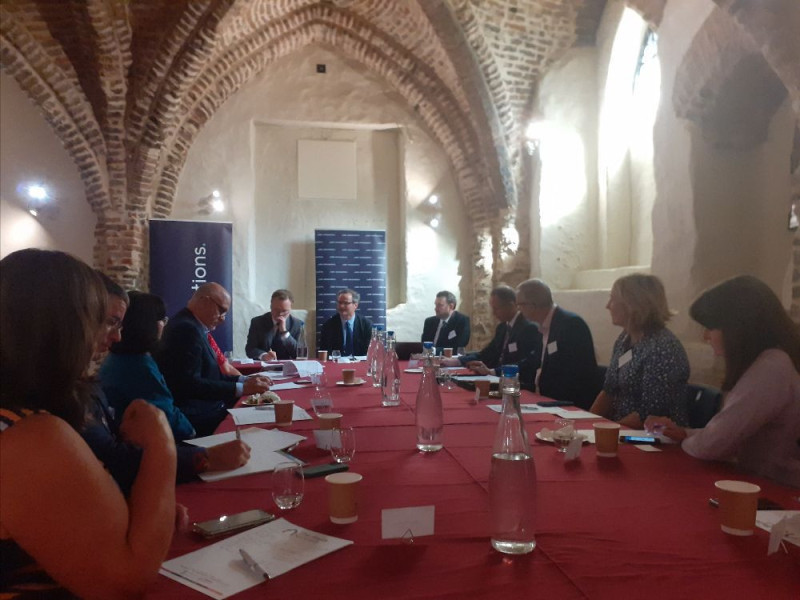 A group of professionals gathered around a long table in a historical venue with brick arches. Various documents and cups are placed on the table. Participants are engaged in discussion, with some taking notes.