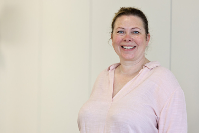 Smiling woman wearing a light pink blouse, standing against a neutral background. Her hair is pulled back, and she looks directly at the camera.