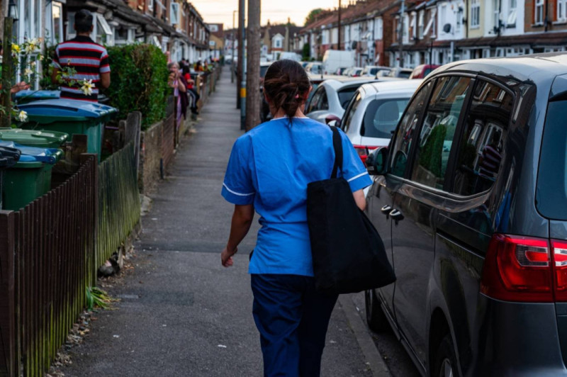 A healthcare worker in blue scrubs walks down a residential street lined with parked cars and houses. There are people visible in the background, some interacting with each other.