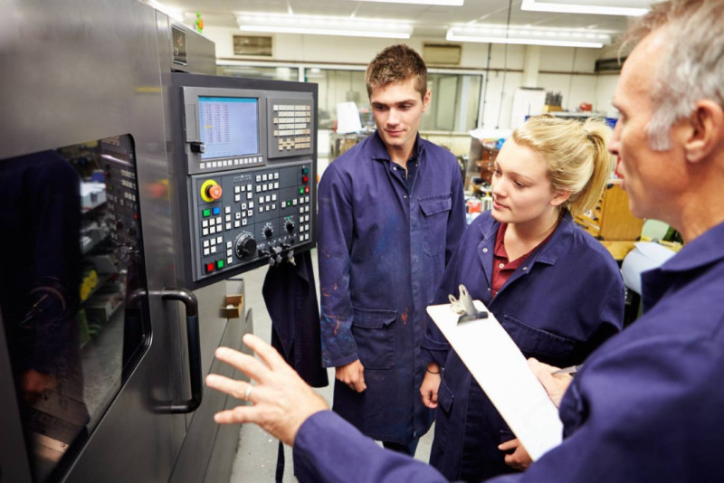 Three people in blue lab coats attentively observe a machinery control panel. One person gestures while explaining to the others, who are listening closely. A clipboard is held by one participant. The setting appears to be an industrial or manufacturing workshop.