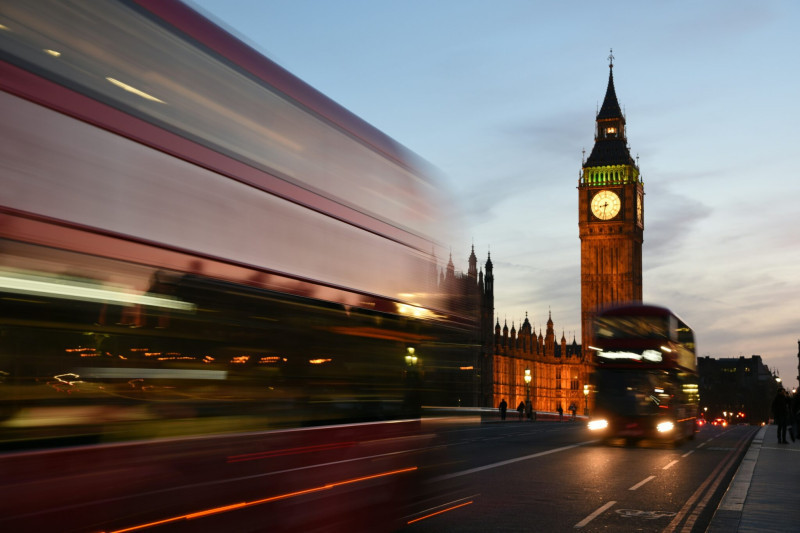 A blurred red double-decker bus passes in front of the iconic Big Ben clock tower during twilight, with warm lights illuminating the nearby buildings.